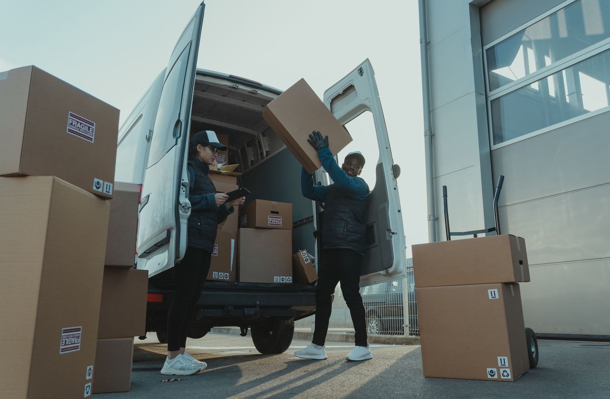 Two logistics team members loading labeled boxes into a delivery van, one holding a tablet