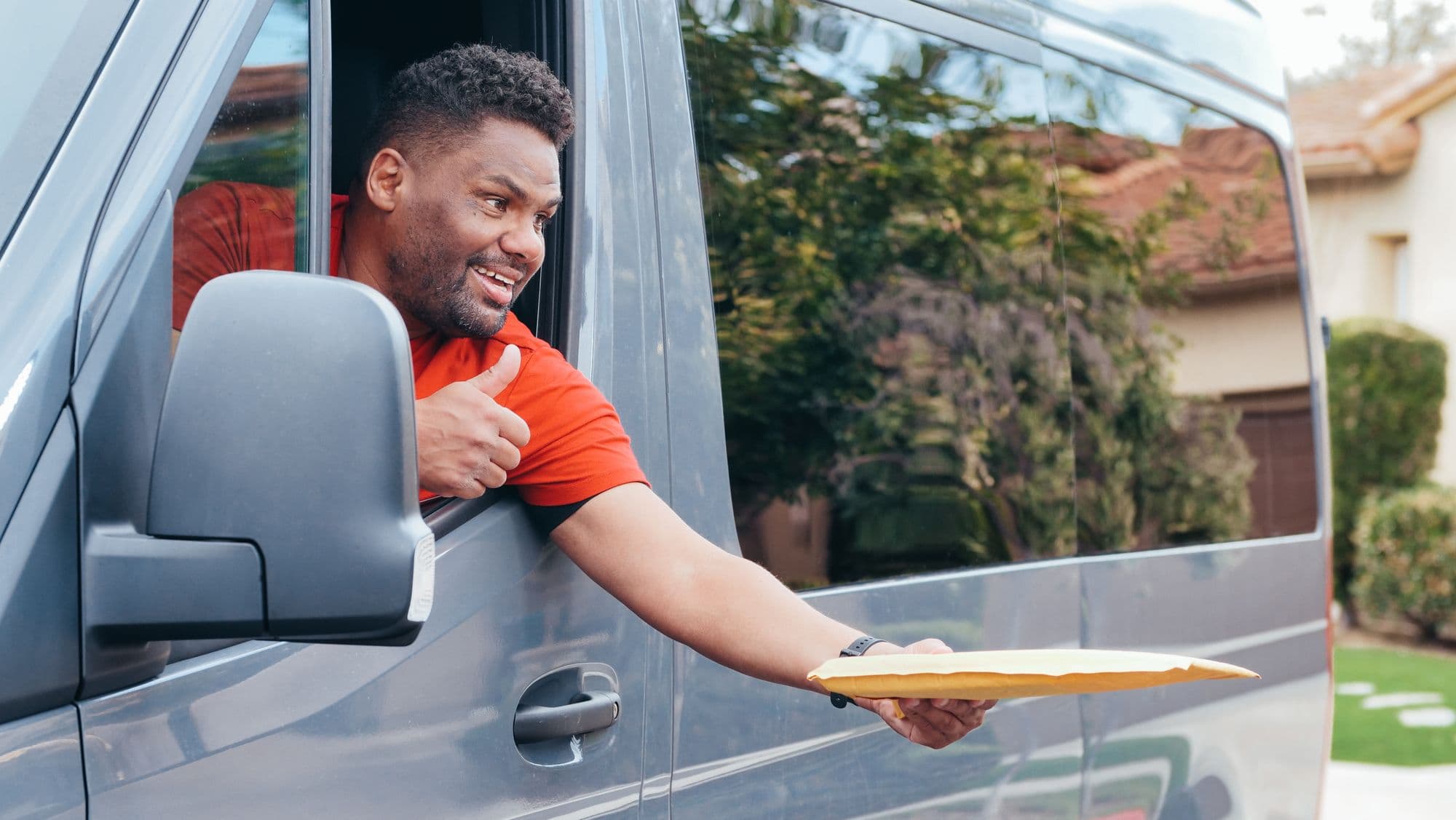 A courier leaning out of an unbranded delivery van window, handing a parcel to a customer